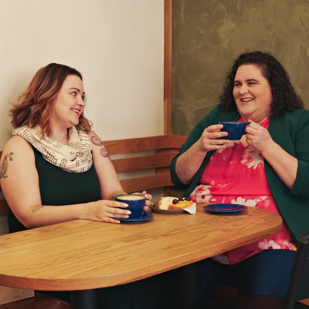 Robyn and Alicia sitting in the corner bench of a coffee shopping, drinking coffee from large blue cups and laughing with eachother.