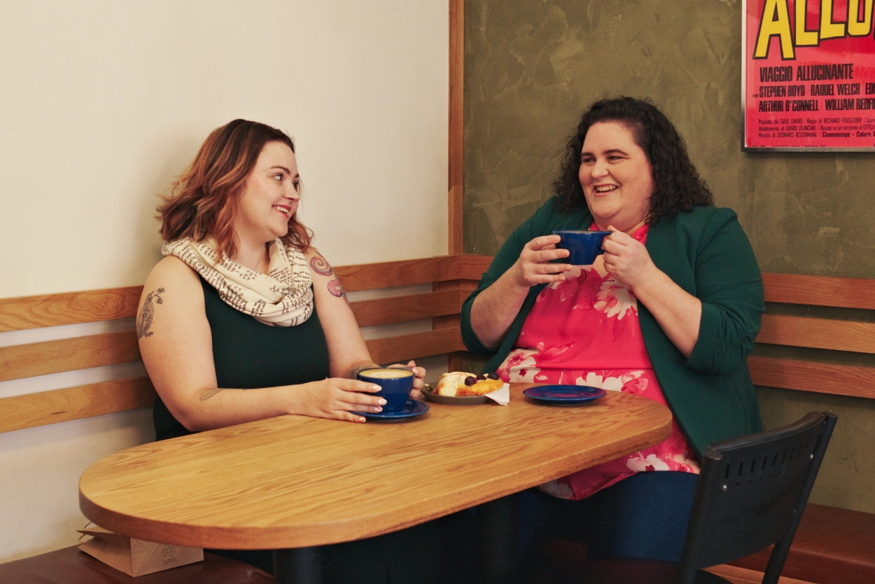 Robyn and Alicia sitting in the corner bench of a coffee shopping, drinking coffee from large blue cups and laughing with eachother.